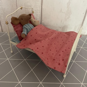 Puede incluir: Una cama de metal blanco en miniatura con una muñeca que lleva una camisa a rayas azules y una cabeza de ratón marrón. La cama tiene una almohada rosa y una manta rosa con lunares rojos. La cama está sobre una superficie con patrón geométrico gris y blanco.