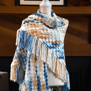 Op de afbeelding: Een gehaakte sjaal in wit, blauw en bruin. De sjaal heeft een franje rand en wordt getoond op een wit mannequin.