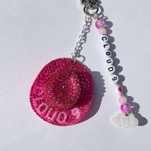 May include: A pink keychain featuring a bedazzled cowboy hat charm. The charm has the text "CLOUD 9" and is accented with a chain, star-shaped clasp, and beaded accents. A white cloud charm completes the design.