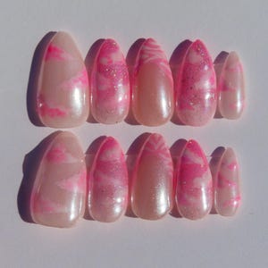 May include: A set of ten almond-shaped false nails. The nails are a pale pink with a marbled design in bright pink. Some nails have glitter accents. The nails are arranged in two rows of five.
