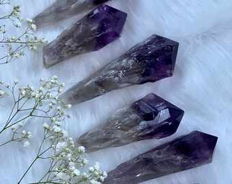 May include: Six amethyst crystal points with a natural, cloudy, and translucent appearance. The points are arranged in a fan shape on a white, fluffy surface.