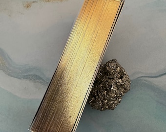 Könnte beinhalten: Ein Stapel goldfarbener Karten liegt auf einer blau-weißen Oberfläche neben einem rauen, dunkelgrauen Stein. Die Karten haben einen metallischen Glanz, und die Kanten sind goldfarben. Der Stein hat eine strukturierte, unebene Oberfläche.