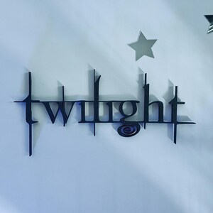 Pode incluir: Um letreiro de parede preto com a palavra twilight em uma fonte estilizada de bordas afiadas com um pequeno detalhe em espiral abaixo da letra i, acompanhado por duas decorações de parede em forma de estrela prateada acima do texto.