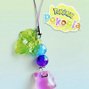 Può includere: Un ciondolo con un personaggio rosa traslucido Ditto con un volto sorridente, collegato a perline blu e viola, una foglia verde e un cinturino viola. Il ciondolo è marchiato con il logo "Pokétopia".