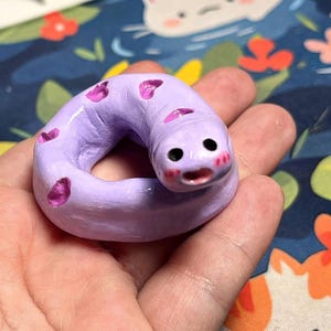 Puede incluir: Una pequeña figura de arcilla color lavanda esculpida a mano con forma de gusano o serpiente enroscada. La figura presenta un cuerpo liso y redondeado con pequeñas manchas rosas hundidas, dos ojos negros circulares y una boca pequeña.