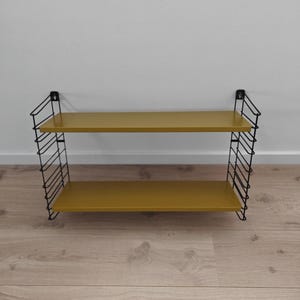 Peut inclure: Étagère murale à deux niveaux avec une surface jaune moutarde et une structure en fil de fer noir. Les étagères sont rectangulaires et le cadre a un design en forme de grille. L'étagère est fixée à un mur blanc.