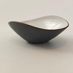 Może przedstawiać: Nowoczesna, ręcznie wykonana ceramiczna miska o unikalnym, asymetrycznym kształcie. Zewnętrzna strona jest teksturowana, ciemnoszara, a wnętrze jasnobiałe. Obręcz ma ciemnobrązowy akcent. Ta dekoracyjna miska jest idealna do serwowania lub ekspozycji.