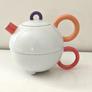 Op de afbeelding: Een witte keramische theepot met een bolvormig lichaam en een deksel. De theepot heeft een rode tuit, een oranje handvat en een rode handgreep aan de zijkant. Het deksel heeft een paarse lusgreep. De theepot staat op drie kleine voetjes.