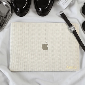 May include: A cream-colored laptop skin with a textured, woven-like pattern and a silver Apple logo in the center. The name Annie is printed in gold lettering in the bottom right corner. The laptop is surrounded by black dress shoes and a watch.