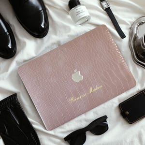 Puede incluir: Una funda para portátil en oro rosa con textura de piel de cocodrilo, con el logotipo de Apple en plata y el texto dorado Primrose Madson, rodeada de zapatos de cuero negro, un frasco de perfume, un reloj de pulsera, una taza de café, guantes y gafas.