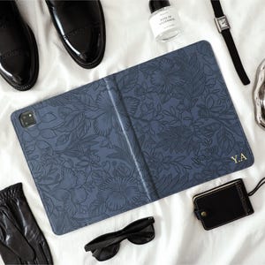 May include: A blue tablet case with a floral design and gold initials "Y.A" is displayed on a white surface, surrounded by black leather gloves, sunglasses, a watch, a perfume bottle, a small black tag, and black leather shoes.