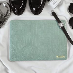 May include: A rectangular laptop sleeve in a light sage green colour with a textured crocodile skin pattern. The bottom right corner features the name Annie printed in gold lettering. The sleeve is surrounded by black dress shoes and a watch.