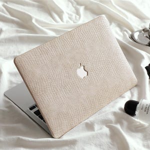 以下が含まれることがあります： 白いシーツの上に置かれた、Appleのロゴが入ったテクスチャード加工のベージュ色のノートパソコン。コーヒーカップ、香水瓶、黒い手袋が入った小さなシルバートレイも近くにあります。