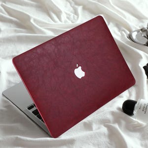 May include: A laptop computer featuring a deep red, textured cover with a prominent white Apple logo in the centre. The device is partially open, revealing a silver metallic base and black keyboard keys, showcasing a sleek and modern aesthetic.