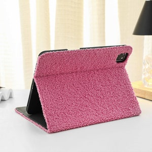 May include: A pink fuzzy case for a tablet or phone. The case is folded to stand upright.