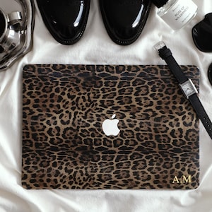May include: A leopard print laptop case with the Apple logo, a black watch, black shoes, and a perfume bottle. The case has the initials "A.M." in gold. The items are arranged on a white surface.