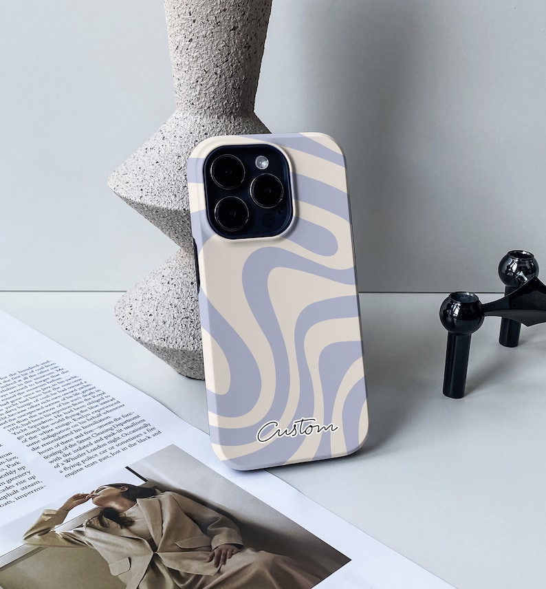 Op de afbeelding: Een lichtblauwe en cr&egrave;me telefoonhoes met een golvend abstract ontwerp. De hoes heeft een uitsparing voor de cameralens en een aangepaste inscriptie.