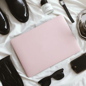 May include: A light pink laptop positioned centrally, surrounded by black leather dress shoes, a small glass perfume bottle labelled Rose of Wilderness, a black wristwatch, a silver tray with a coffee cup, a black leather card holder, and black sunglasses.