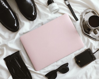 Op de afbeelding: Een lichtroze laptop in het midden, omringd door zwarte leren nette schoenen, een klein glazen parfumflesje met het label Rose of Wilderness, een zwart polshorloge, een zilveren dienblad met een koffiekopje, een zwarte leren kaarthouder en een zwarte zonnebril.