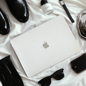 May include: A white textured laptop skin with a silver Apple logo and gold monogram letters, surrounded by black leather dress shoes, a perfume bottle, a wristwatch, a silver cup, a black leather card holder, and black sunglasses.