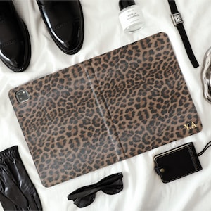 Può includere: Una custodia per tablet con stampa leopardata marrone e nera con le iniziali dorate "Y.A". L'immagine mostra anche guanti in pelle nera, occhiali da sole, un orologio, un flacone di profumo, un vassoio d'argento con una tazza e scarpe in pelle nera.