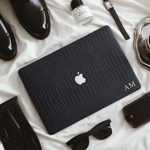 May include: A flat lay featuring a black laptop case with a textured, crocodile-like pattern and the Apple logo. Other items include black leather shoes, sunglasses, a watch, a perfume bottle, a cup of coffee, a silver tray, a leather tag, and black gloves.