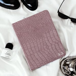 May include: A mauve faux-crocodile-skin journal with the initials "K.S" embossed on the cover. The journal is surrounded by black dress shoes, sunglasses, a perfume bottle, and a silver tray, all set on a white surface.