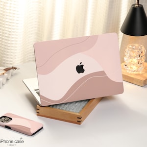 以下が含まれることがあります： アップルのロゴが付いたピンクとブラウンの抽象的なデザインのラップトップスキン。スキンは閉じたラップトップコンピューターの上にあります。ラップトップは木製の箱の上に置かれています。ピンクとブラウンのiPhoneケースが手前にあります。