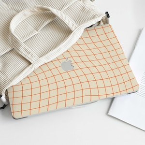 May include: A beige corduroy tote bag with a laptop case featuring a grid pattern in orange. The laptop case has a silver Apple logo. A magazine with the text "CATCH THE" is next to the bag.