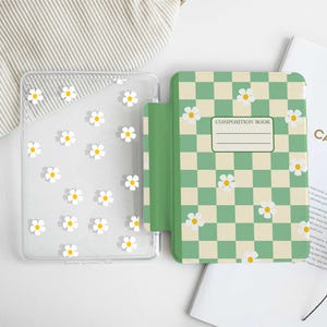 Op de afbeelding: Een doorzichtige telefoonhoes met witte madeliefjesaccenten naast een groen en crème geruit compositieboek, eveneens met madeliefjesaccenten. Het compositieboek heeft een label met de tekst "COMPOSITION BOOK".