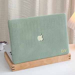 Könnte beinhalten: Hellgrüne Laptop-Hülle mit Krokodilhaut-Textur. Die Hülle hat ein goldenes Apple-Logo und goldene Initialen "D.V." unten rechts. Der Laptop steht auf einem Holzständer.