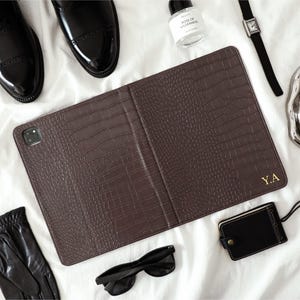 May include: A brown, crocodile-textured leather tablet case, monogrammed with the gold initials "Y.A.". The image also features black leather gloves, sunglasses, a watch, and black dress shoes, all displayed on a white background.