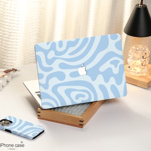K&ouml;nnte beinhalten: Eine hellblaue Laptop-Haut mit einem wei&szlig;en abstrakten Wirbelmuster. Die Haut befindet sich auf einem Laptop mit einem silbernen Apple-Logo. Der Laptop steht auf einer Holzoberfl&auml;che. Auf der Oberfl&auml;che befindet sich eine hellblaue iPhone-H&uuml;lle mit dem gleichen Muster.