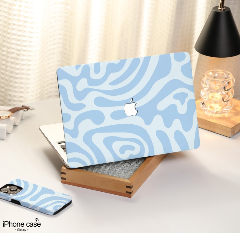 K&ouml;nnte beinhalten: Eine hellblaue Laptop-Haut mit einem wei&szlig;en abstrakten Wirbelmuster. Die Haut befindet sich auf einem Laptop mit einem silbernen Apple-Logo. Der Laptop steht auf einer Holzoberfl&auml;che. Auf der Oberfl&auml;che befindet sich eine hellblaue iPhone-H&uuml;lle mit dem gleichen Muster.