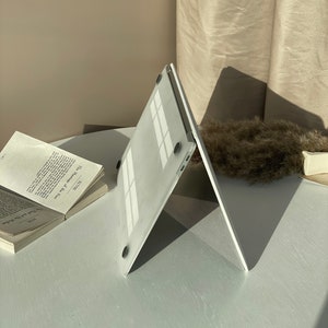 Op de afbeelding: Een witte laptophoes met een textuur en zwarte rubberen voetjes. De hoes is open en in een hoek gekanteld.