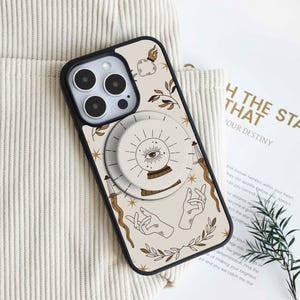 May include: A phone case with a black border and a cream-colored background featuring a mystical design. The design includes an eye, hands, stars, and a pop-up grip. The pop-up grip has a circular design with an eye and sunburst pattern.