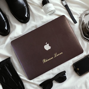 May include: A dark brown laptop skin featuring a white Apple logo and the gold cursive text Rhianne Lioran. The laptop is surrounded by black leather dress shoes, a perfume bottle, a wristwatch, a coffee cup, sunglasses, and a leather tag.