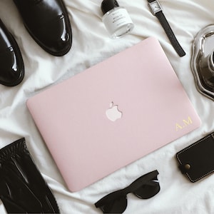 May include: A flat lay featuring a pink laptop with the Apple logo and gold initials, black sunglasses, a black leather wallet, a silver tray with a cup of coffee, a bottle of perfume, a watch, black leather gloves, and black leather shoes.