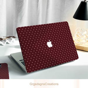 Könnte beinhalten: Eine weinrote Laptop-Hülle mit weißem Apple-Logo und einem Muster aus kleinen, gleichmäßig verteilten, hellrosa Punkten. Der Laptop ist auf einem weißen Schreibtisch geöffnet. Der Text "GrgsdsgnsCreations" befindet sich unten.