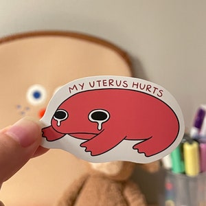 Puede incluir: Una pegatina de rana roja con una expresi&oacute;n triste y el texto "MY UTERUS HURTS" sobre un fondo blanco.