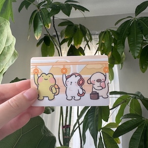 May include: A rectangular sticker featuring three cartoon characters: a yellow frog, a white creature with headphones, and a beige dog with a suitcase. The characters are in a train car setting with orange handrails and a light beige background.