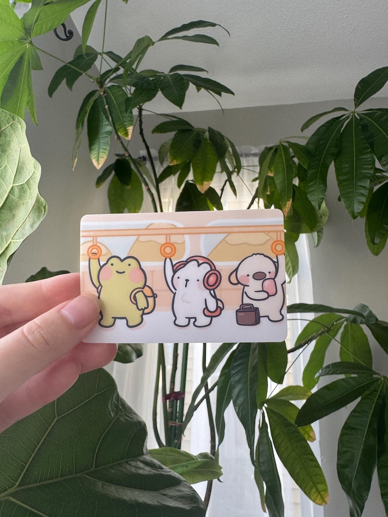 May include: A rectangular sticker featuring three cartoon characters: a yellow frog, a white creature with headphones, and a beige dog with a suitcase. The characters are in a train car setting with orange handrails and a light beige background.