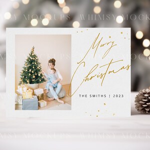 May include: A white Christmas card with a gold inscription that reads "Merry Christmas" and "The Smiths | 2023". The card features a photo of a mother and child decorating a Christmas tree.
