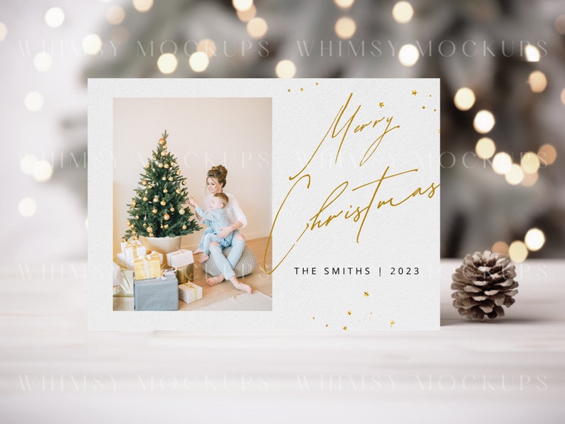 May include: A white Christmas card with a gold inscription that reads "Merry Christmas" and "The Smiths | 2023". The card features a photo of a mother and child decorating a Christmas tree.