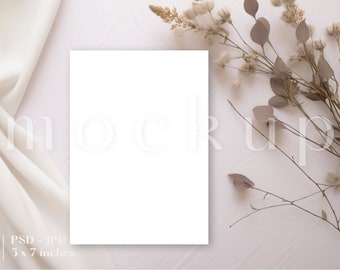 May include: A white card mockup with a blank white card on a white background with dried flowers and a white fabric.