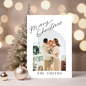 May include: A white Christmas card with a photo of a family of three. The card has a gold arch and the text "Merry Christmas" in black cursive. The family is standing in front of a Christmas tree and the text "The Smiths" is at the bottom of the card.