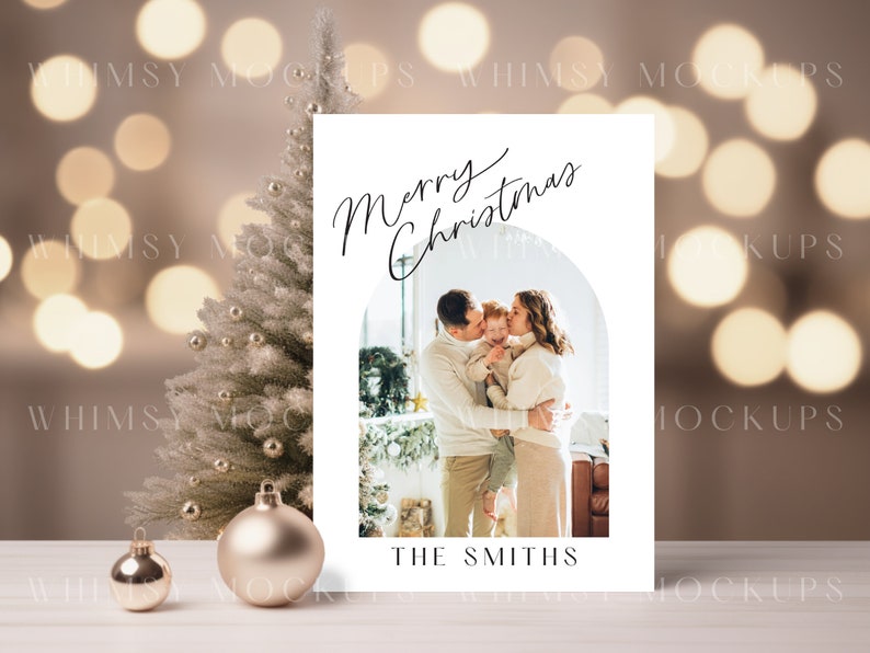 May include: A white Christmas card with a photo of a family of three. The card has a gold arch and the text "Merry Christmas" in black cursive. The family is standing in front of a Christmas tree and the text "The Smiths" is at the bottom of the card.