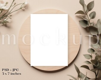 Puede incluir: Una maqueta de tarjeta blanca en blanco sobre una superficie redonda de madera con follaje verde y flores blancas sobre un fondo beige. La tarjeta mide 12,7 x 17,8 cm.