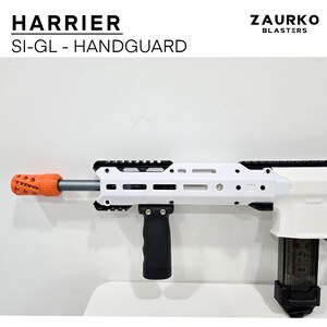 Puede incluir: Un guardamanos Harrier SI-GL blanco y negro con un freno de boca naranja. El guardamanos est&aacute; unido a un blaster blanco con una empu&ntilde;adura vertical negra. El texto "Zaurko Blasters" es visible en la esquina superior derecha de la imagen.