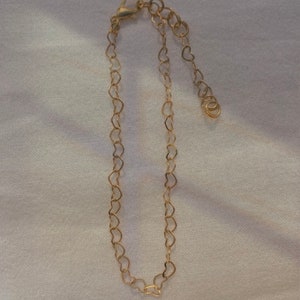 Puede incluir: Una delicada pulsera dorada con eslabones en forma de corazón. La pulsera tiene un cierre de mosquetón y una cadena ajustable. La joya se muestra sobre una superficie blanca y texturizada.